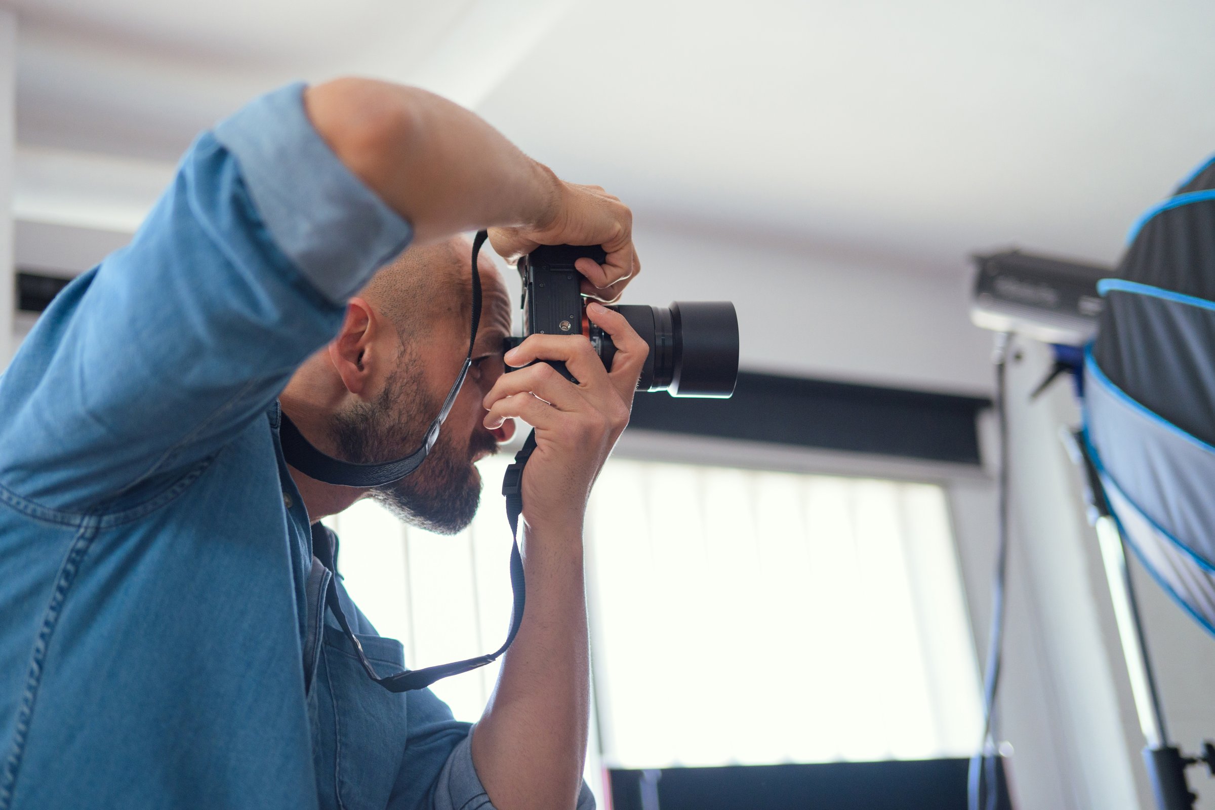 A photographer shoots a female model in a studio. The model is standing, wearing a black top and blue jeans. The photographer is holding a camera. The background is a black backdrop a with softbox