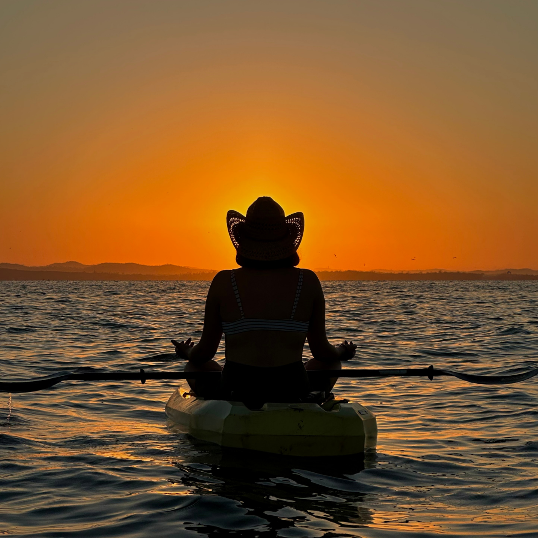 Silhouette of a person in a hat meditating on a kayak during a sunset over calm waters.