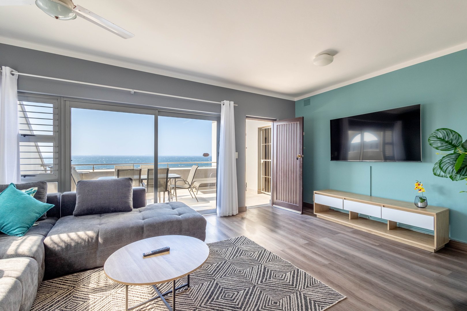 Modern living room with a gray sofa, flat-screen TV, large window, and balcony view of the ocean.
