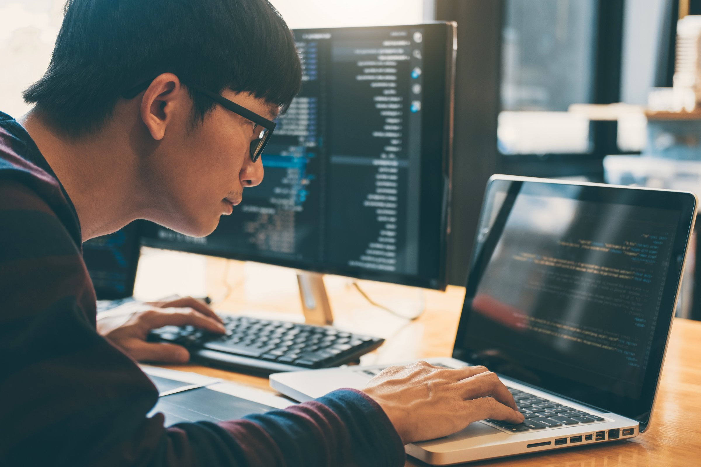 Person working on a laptop with code on screen, seated at a desk with two additional monitors displaying text.