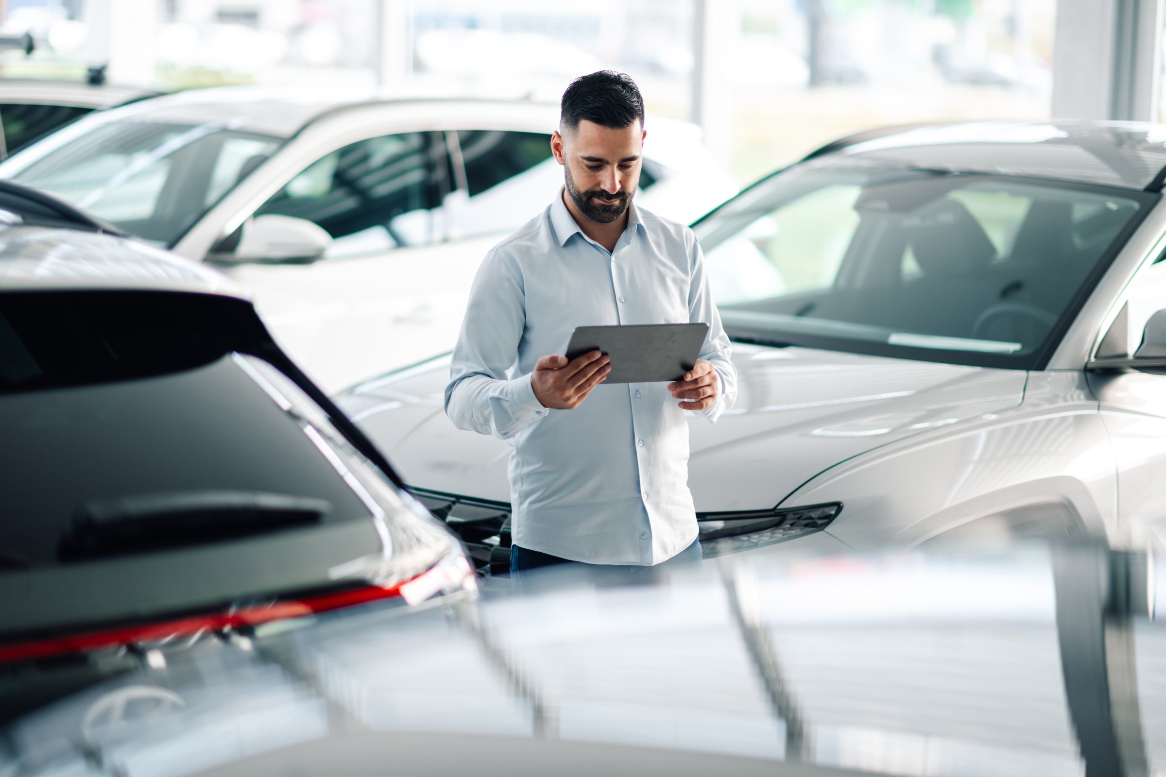 Focused car salesman in auto showroom uses digital tablet, showcasing professionalism. Modern setting and wireless tech add to business atmosphere as he assists customers in choosing vehicles