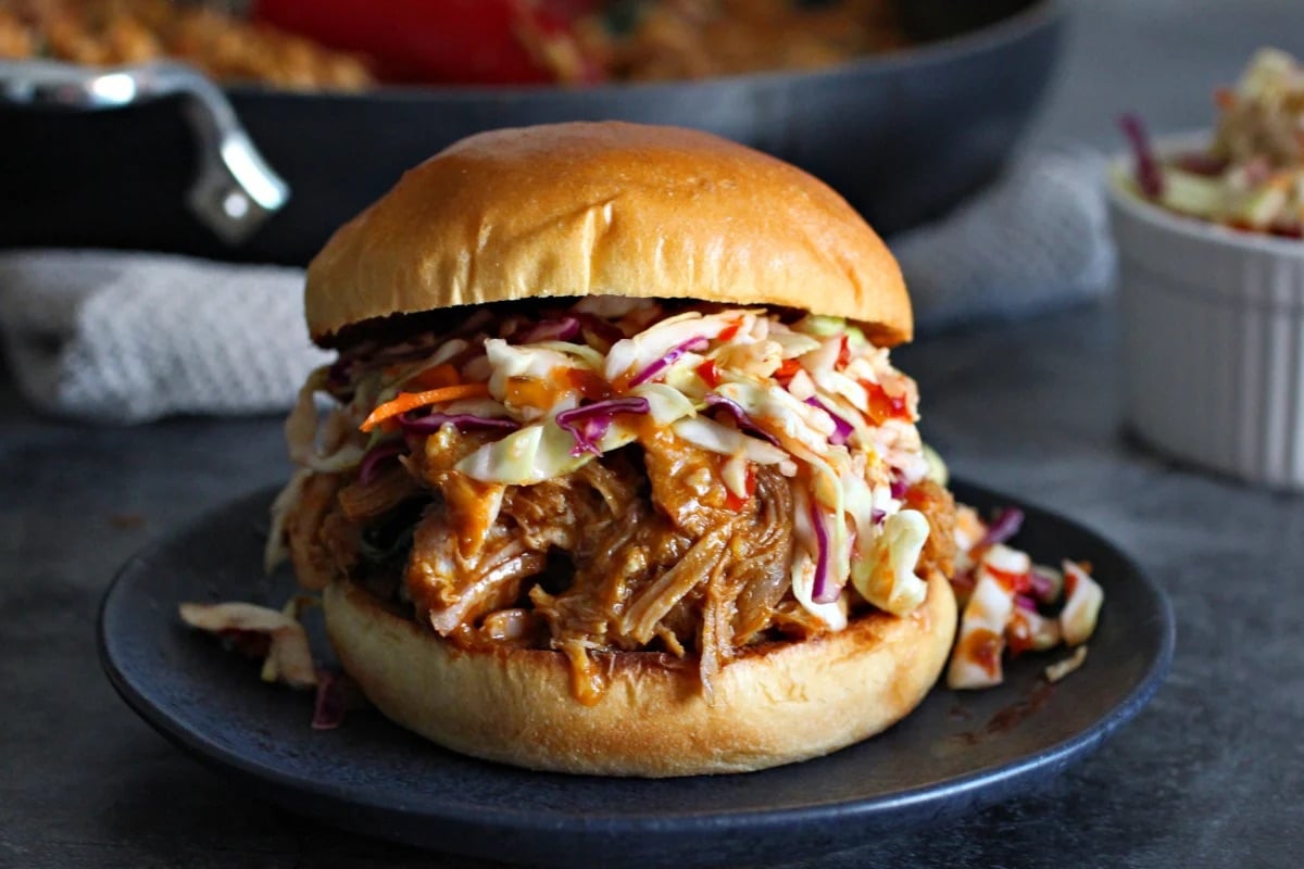 Pulled pork sandwich with coleslaw on a bun, served on a dark plate beside a small side of coleslaw.