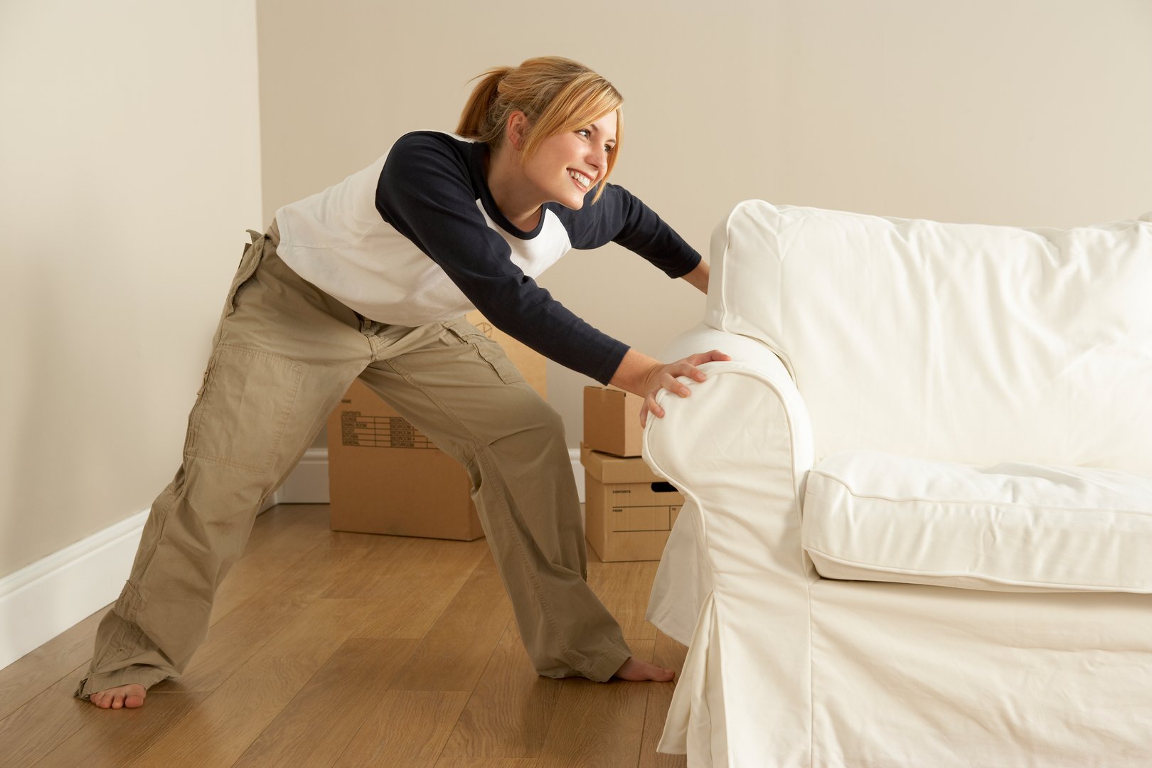 Woman in casual clothing smiling while pushing a white sofa in a room with cardboard boxes on a wooden floor.