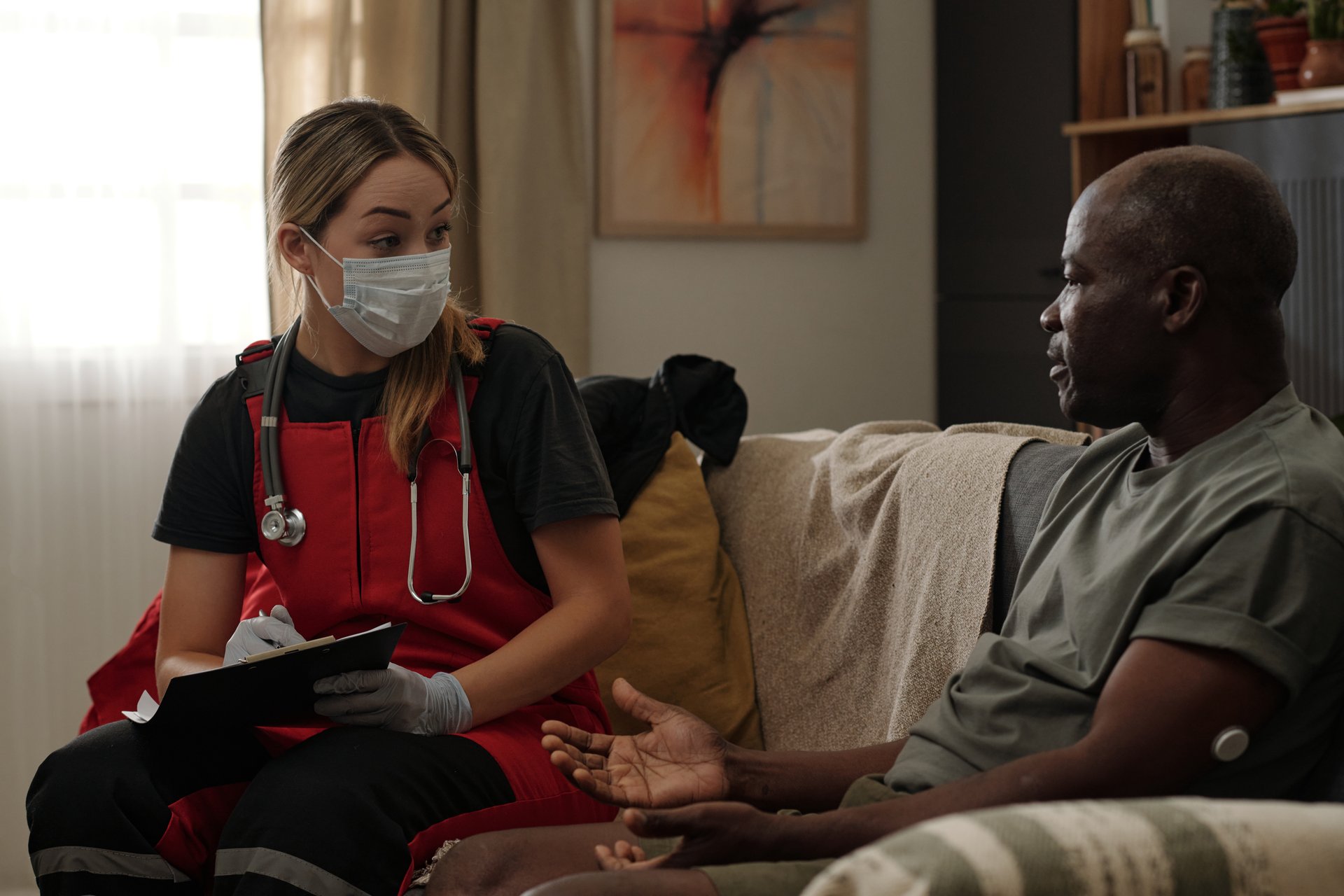 Healthcare professional in uniform speaking with senior patient sitting on couch, holding clipboard and taking notes while both are wearing masks following health protocols
