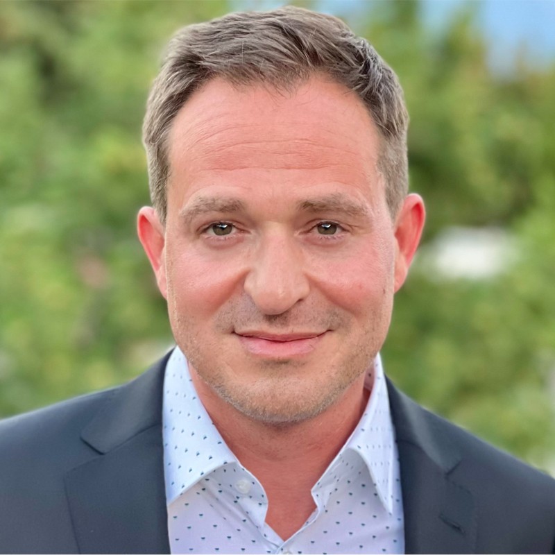 Smiling man in a suit with a light blue shirt, standing outdoors with a blurred green background.