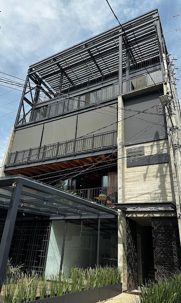 Modern multi-story building with metal and glass facade, featuring balconies and a sign by the entrance, under a cloudy sky.