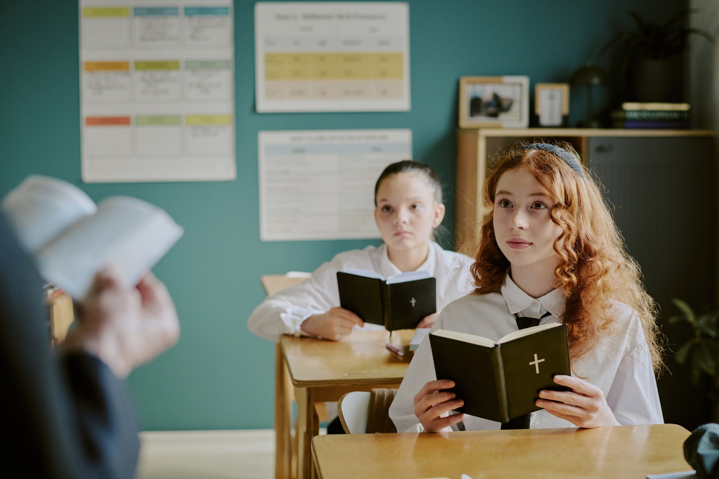 Portrait of two students sitting at desks looking at teacher reading holy book in foreground, modern blue green wall with charts in background