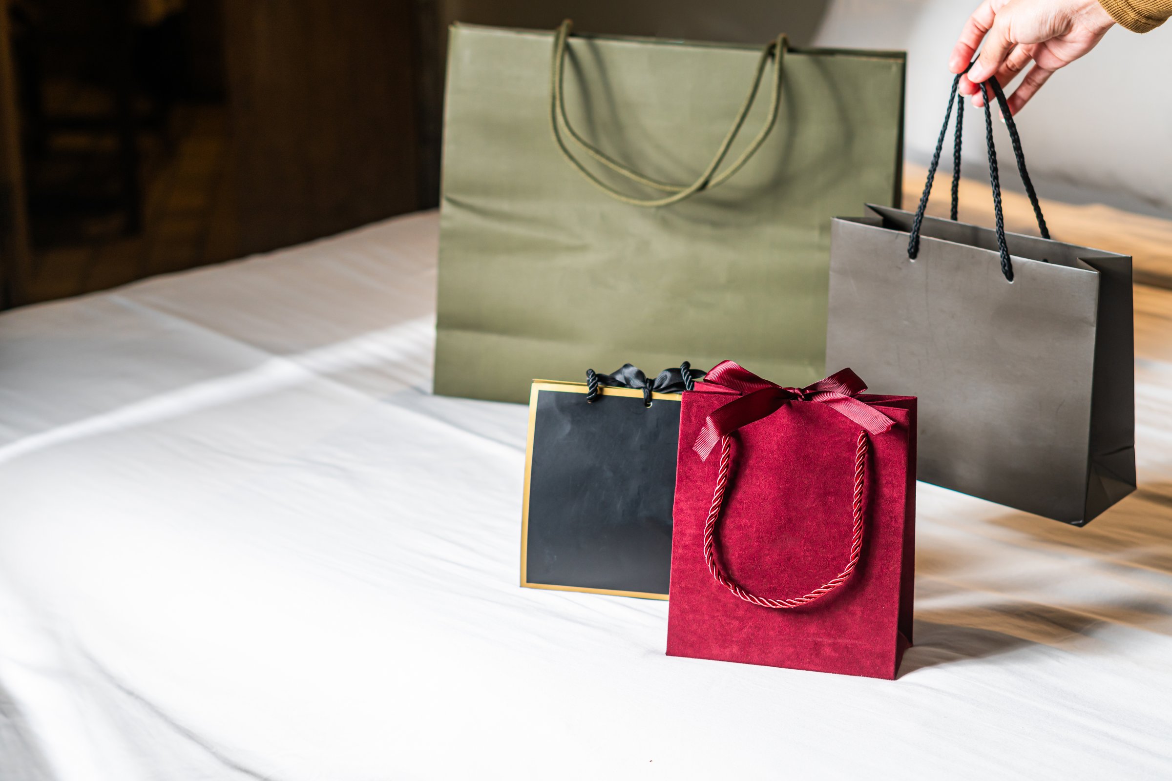A collection of shopping bags in various sizes and colors placed on a bed. The bags include a large green bag, a medium black bag, and a small red bag with a bow. The setting is cozy and inviting