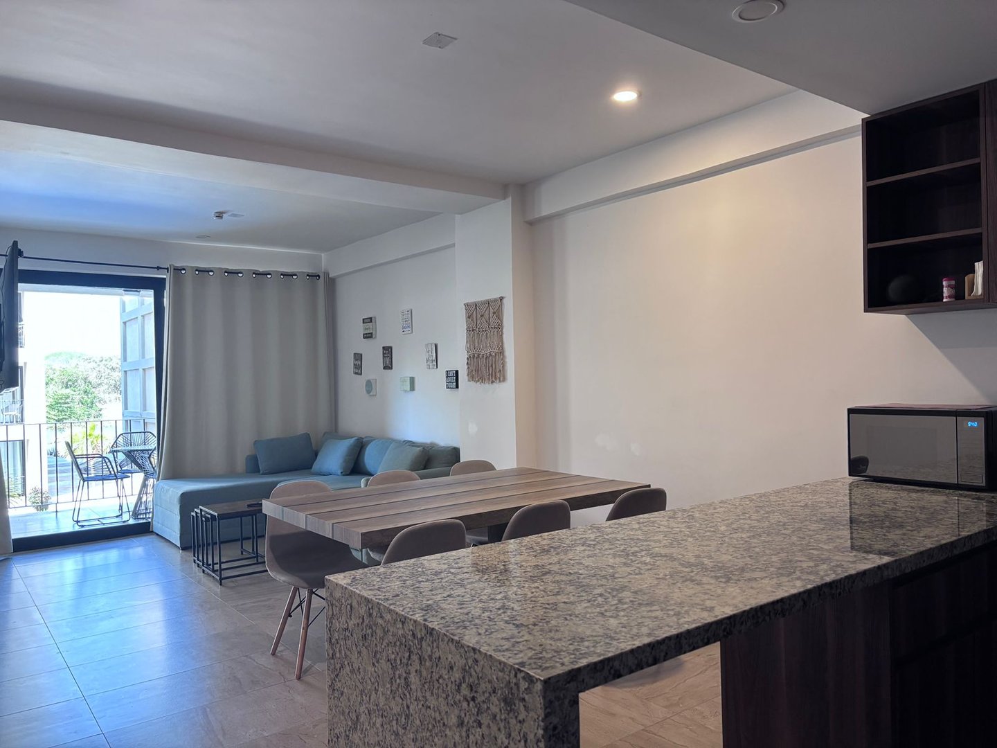 Modern living room with a sectional sofa, dining table, balcony view, and a kitchen counter with a microwave.