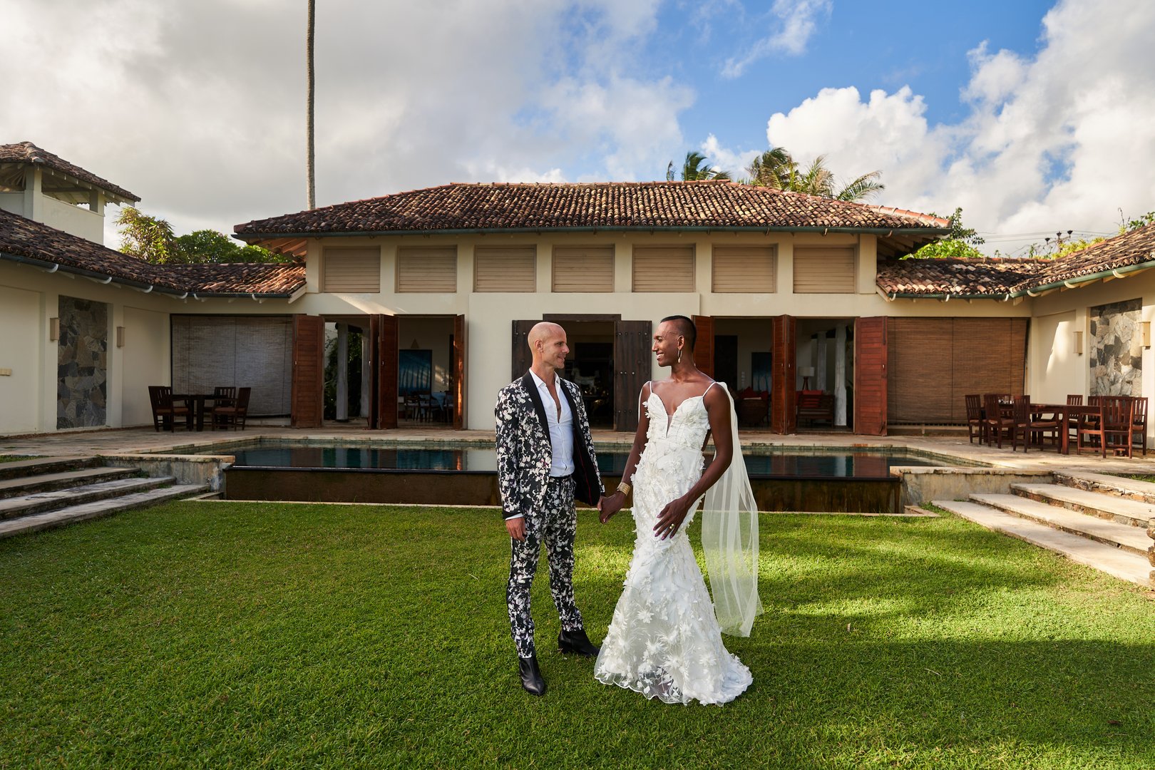 Gender fluid bride in white gown holds hand with groom in patterned suit at tropical villa wedding. LGBTQ couple enjoy vibrant ceremony, with rich plants. Celebrate love and diversity.