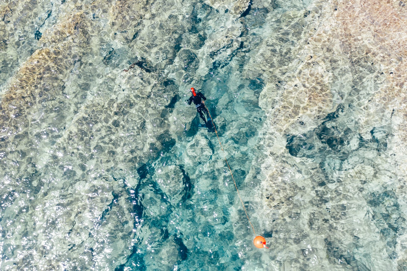 High angle view of man spearfishing in shallow water. Drone view of spearfisherman in wetsuit with buoy and speargun looking for a fish.