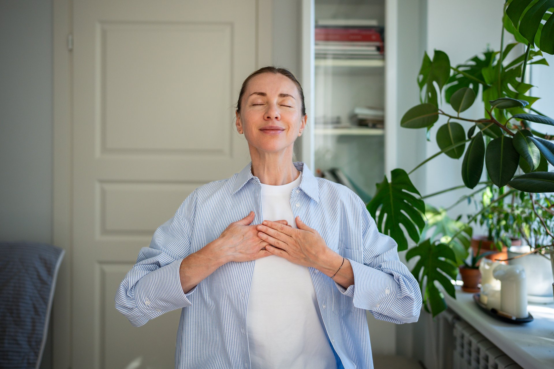 Relaxed smiling woman meditates with hands on chest, practicing breathing exercises at home demonstrating mindfulness techniques to cope with emotional eating