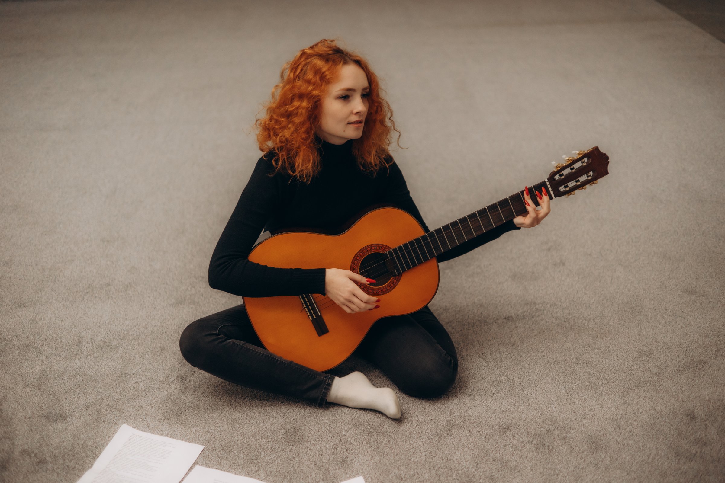 A red-haired woman sits on a carpet, strumming an acoustic guitar, surrounded by papers, evoking a calm and creative atmosphere in an indoor setting.