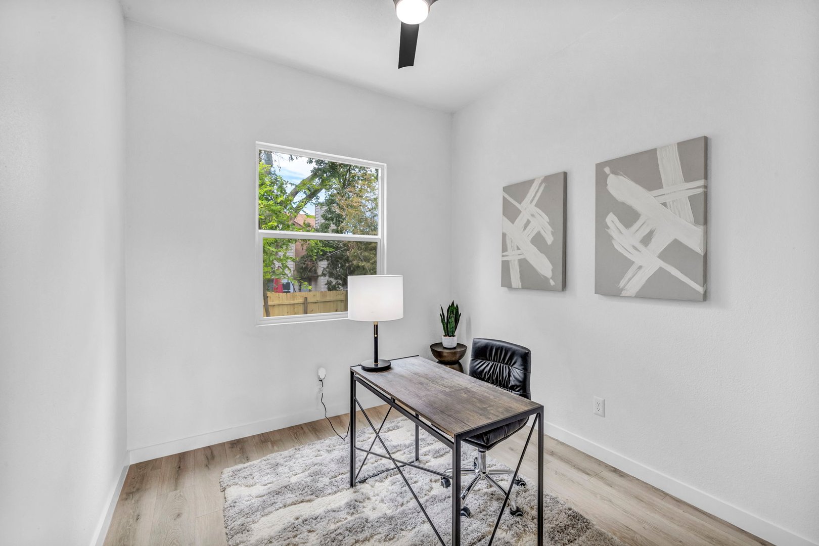 Minimalist office with desk, chair, lamp, and abstract art on the walls. Large window and light wood flooring.