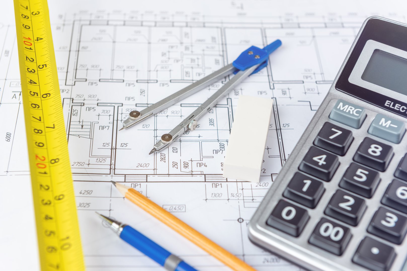 An architect's workspace with blueprints, a calculator, a measuring tape, a compass, a pencil, and an eraser symbolizes precision, planning, and engineering in construction and architectural design projects.