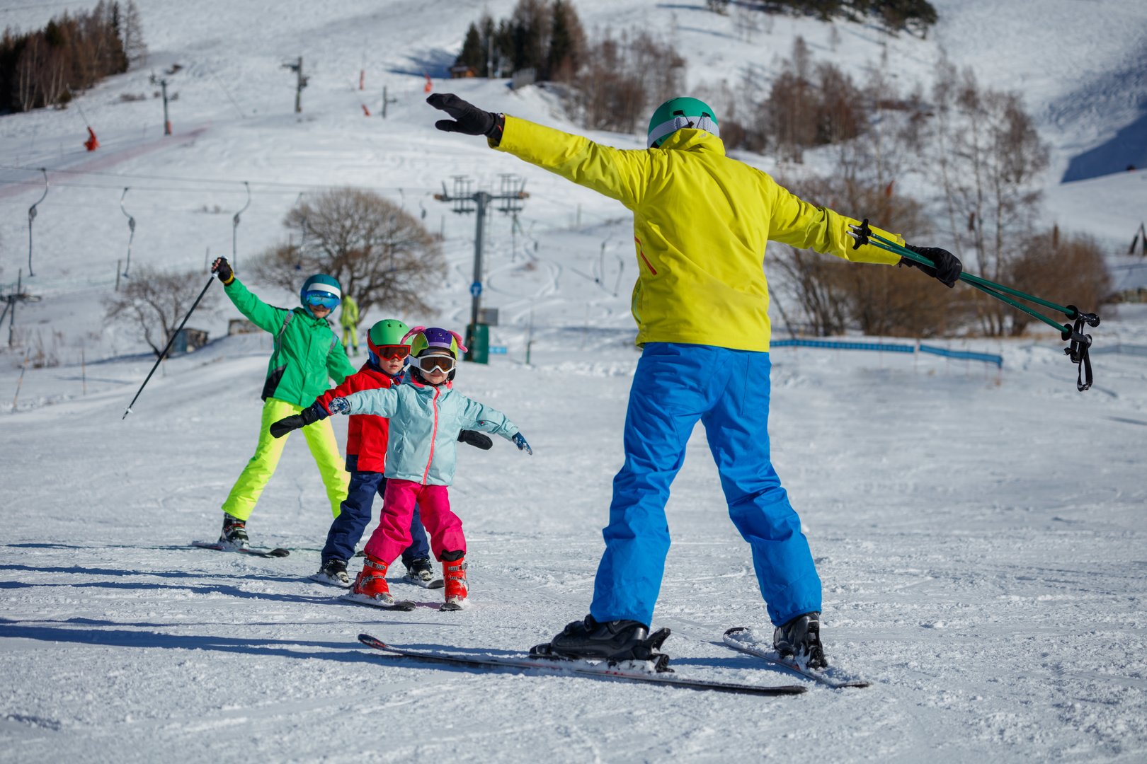 An instructor on a snowy mountain demonstrates skiing techniques to three children, all dressed in eye-catching winter gear on alpine resort