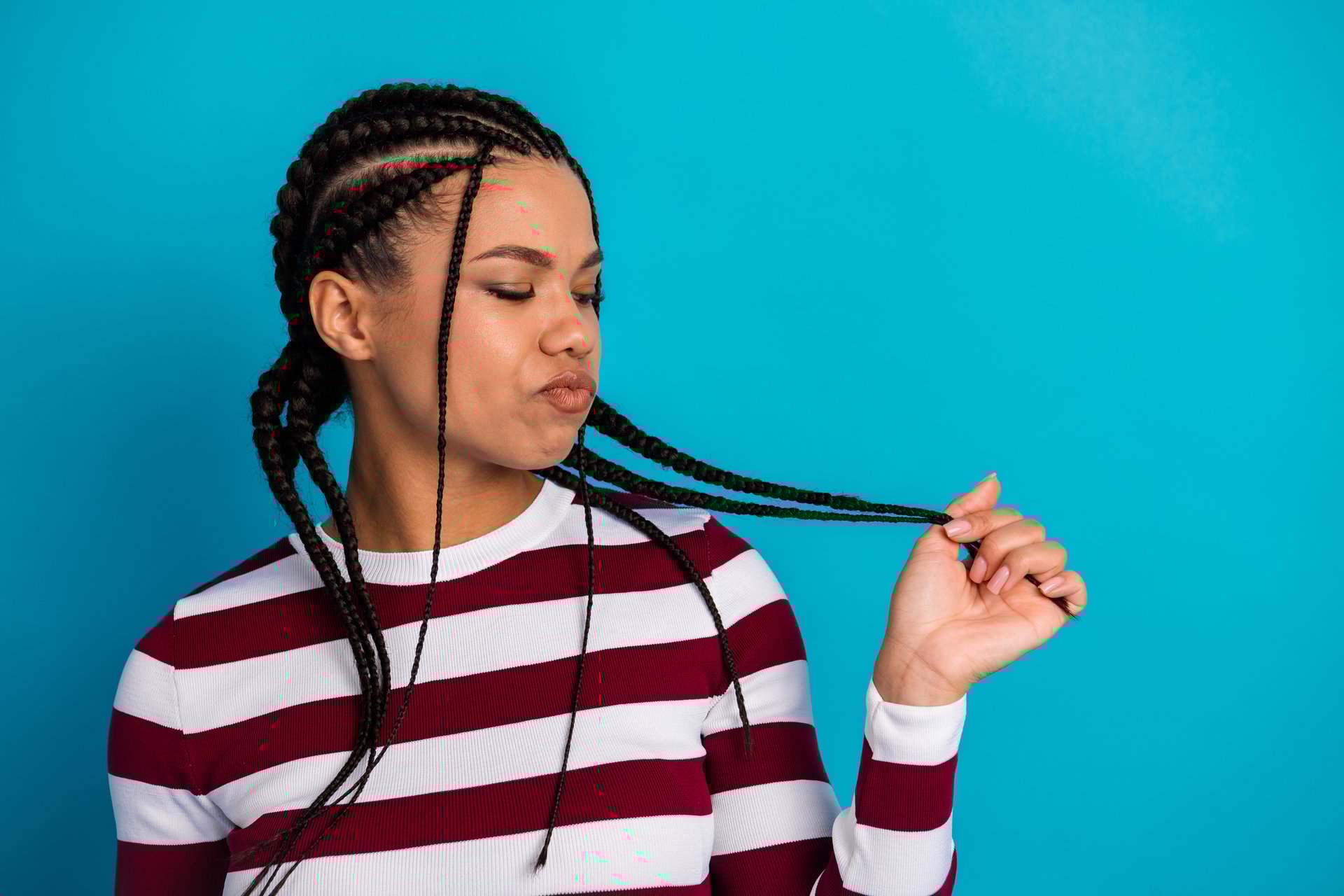 A stylish portrait of a young woman with cornrow braids wearing a striped maroon and white top, tilting her head while playing with her hair against a vibrant blue backdrop. Playful confident and fashionable vibe.