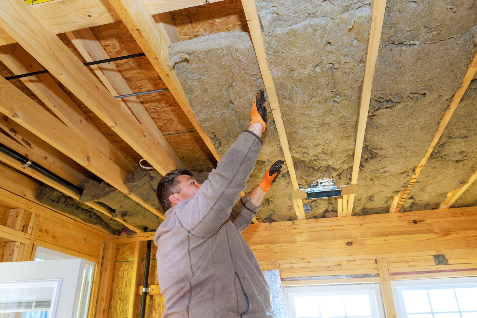 Worker is fitting insulation material into wooden ceiling beams of house, enhancing energy efficiency.