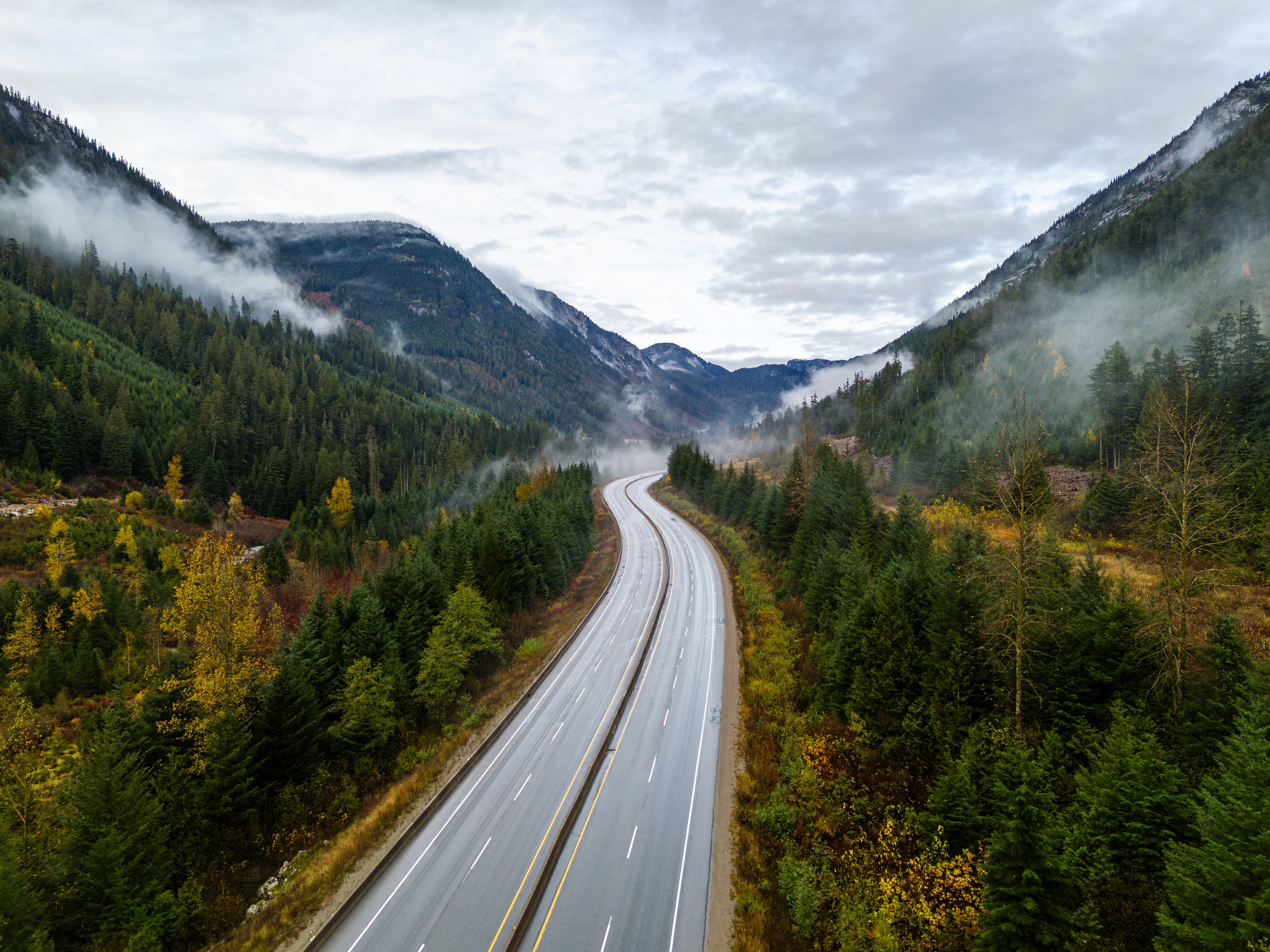 A breathtaking aerial view of the Coquihalla Highway winding through lush forests and misty mountains in British Columbia, Canada. The scene captures the serene beauty of nature and landscape.
