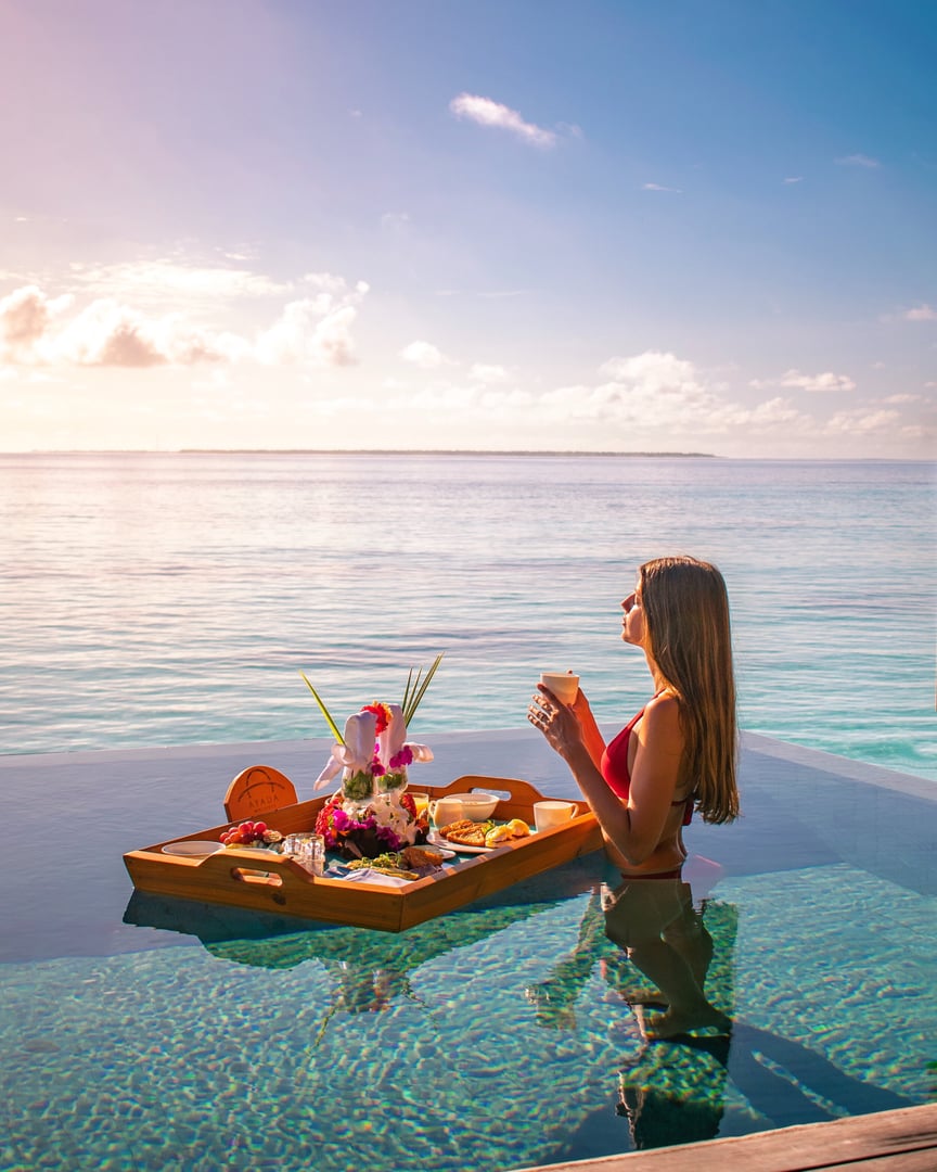 Woman in a swimsuit enjoys breakfast on a floating tray in an infinity pool overlooking the ocean.