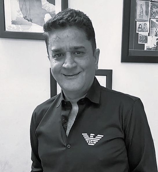 A man smiling, wearing a black shirt with an Armani logo, standing in front of framed pictures on the wall.