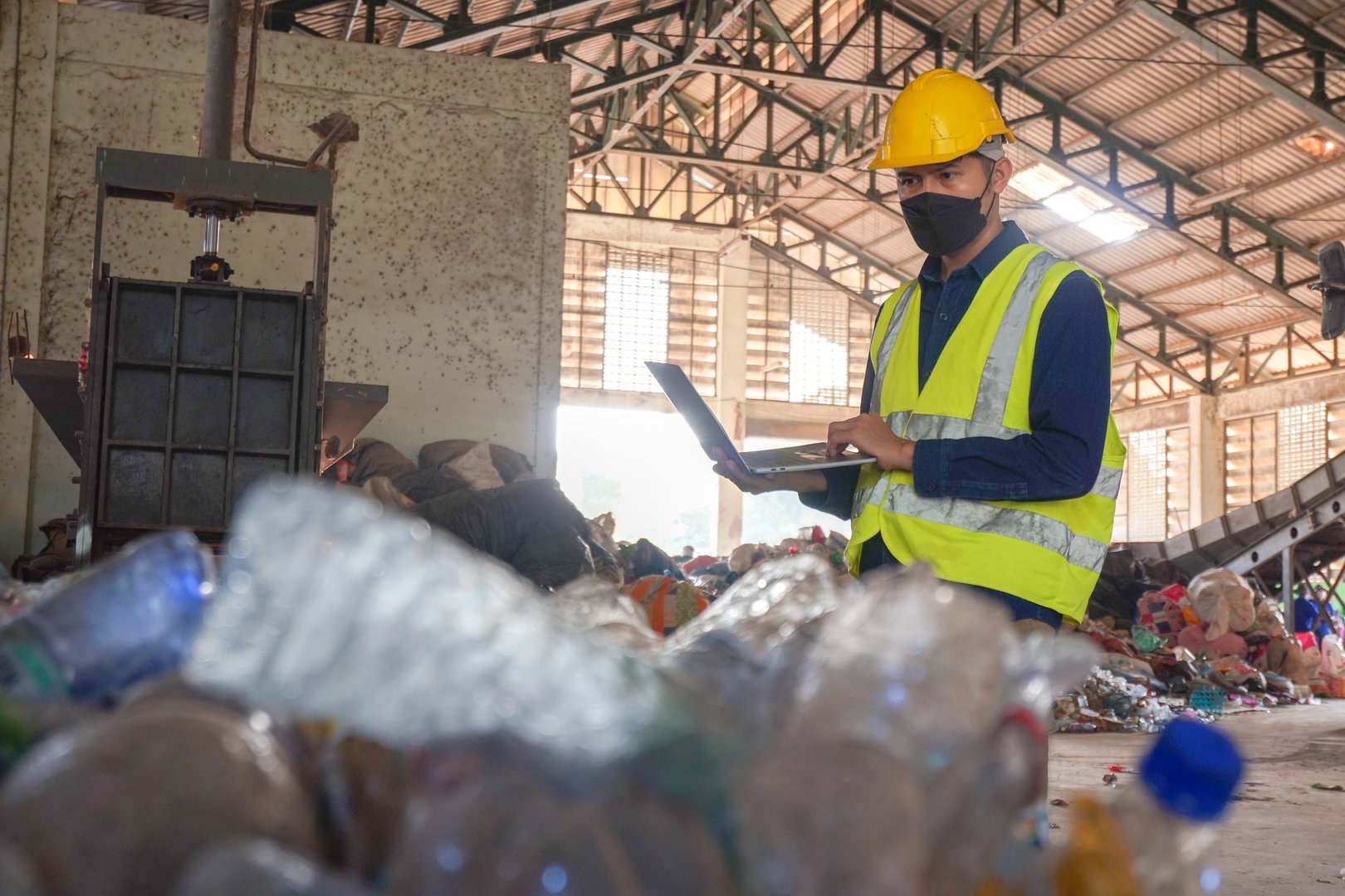 Asian environmental engineer uses laptop to check quality of compressed waste plastic for transportation or production in power industry at waste separation plant.