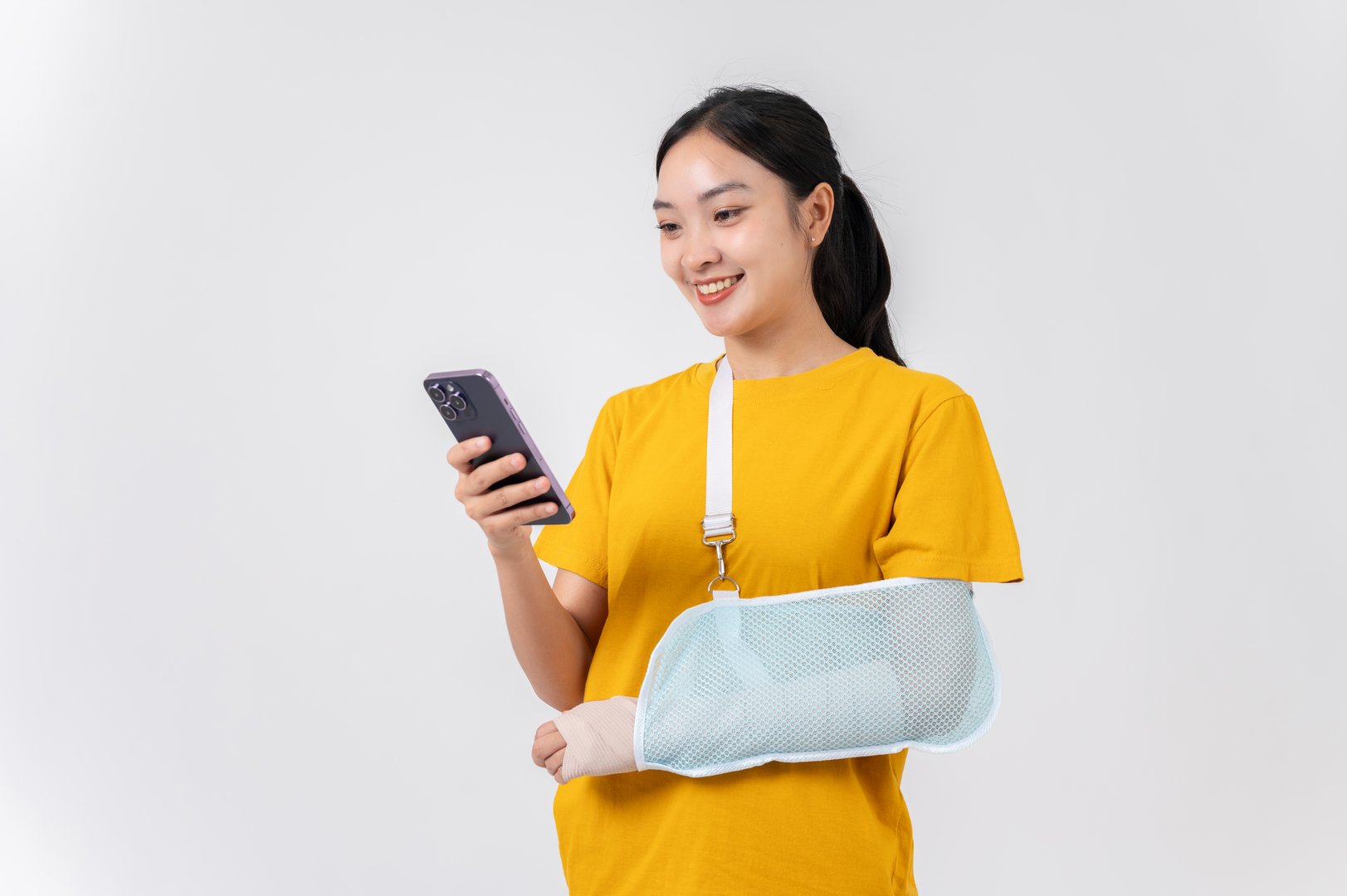 A smiling, carefree Asian woman with her broken arm, wrapped in a plaster bandage and supported by a sling after an accident injury, using her smartphone. health care and life insurance