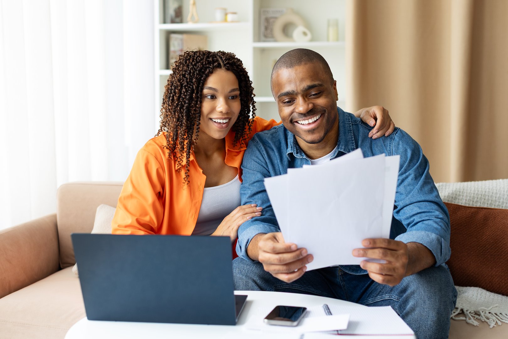 Happy young African American couple reviewing paperwork together at home, checking documents, smiling and sitting at table with laptop in living room