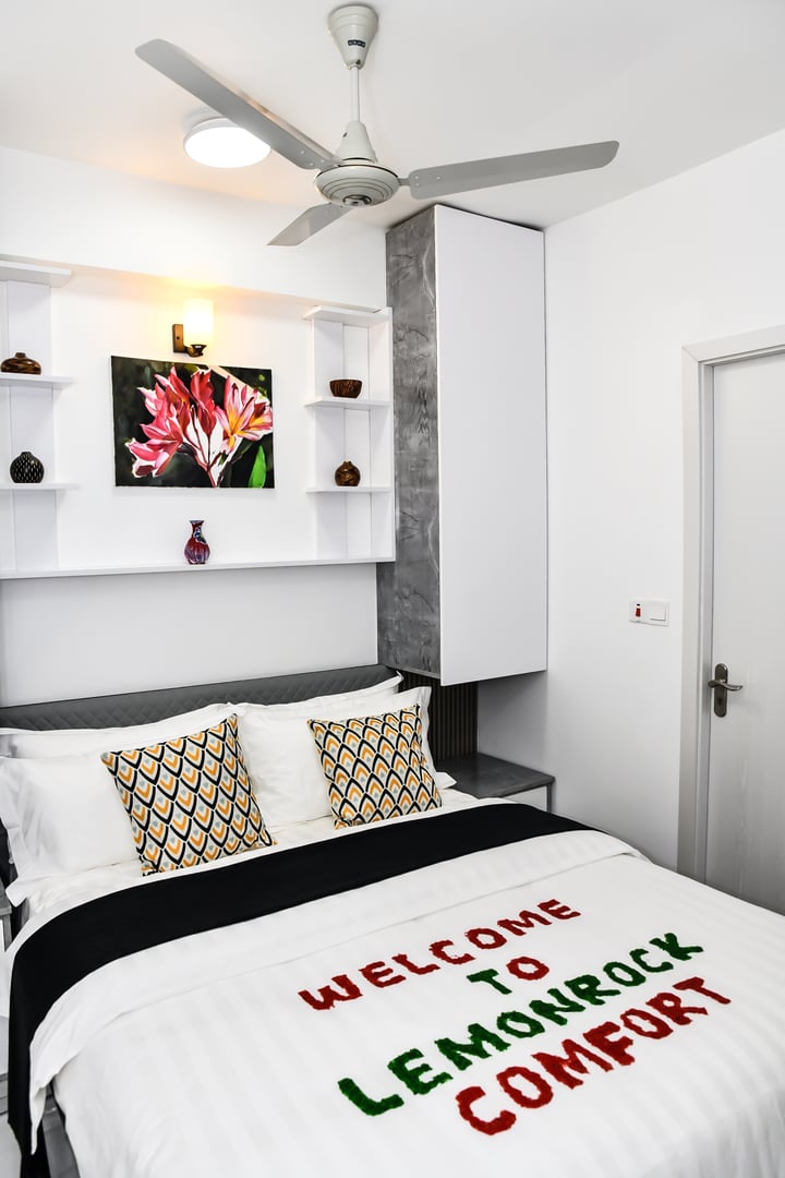 Modern bedroom with a bed featuring a Welcome to Lemonrock Comfort message on the duvet, art piece on shelf, and ceiling fan.