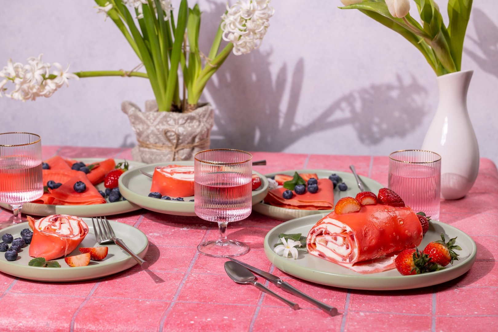 Pink strawberry dessert crepes and rolls served on a table decorated with flowers and glasses of water, creating a stylish and delicious setting