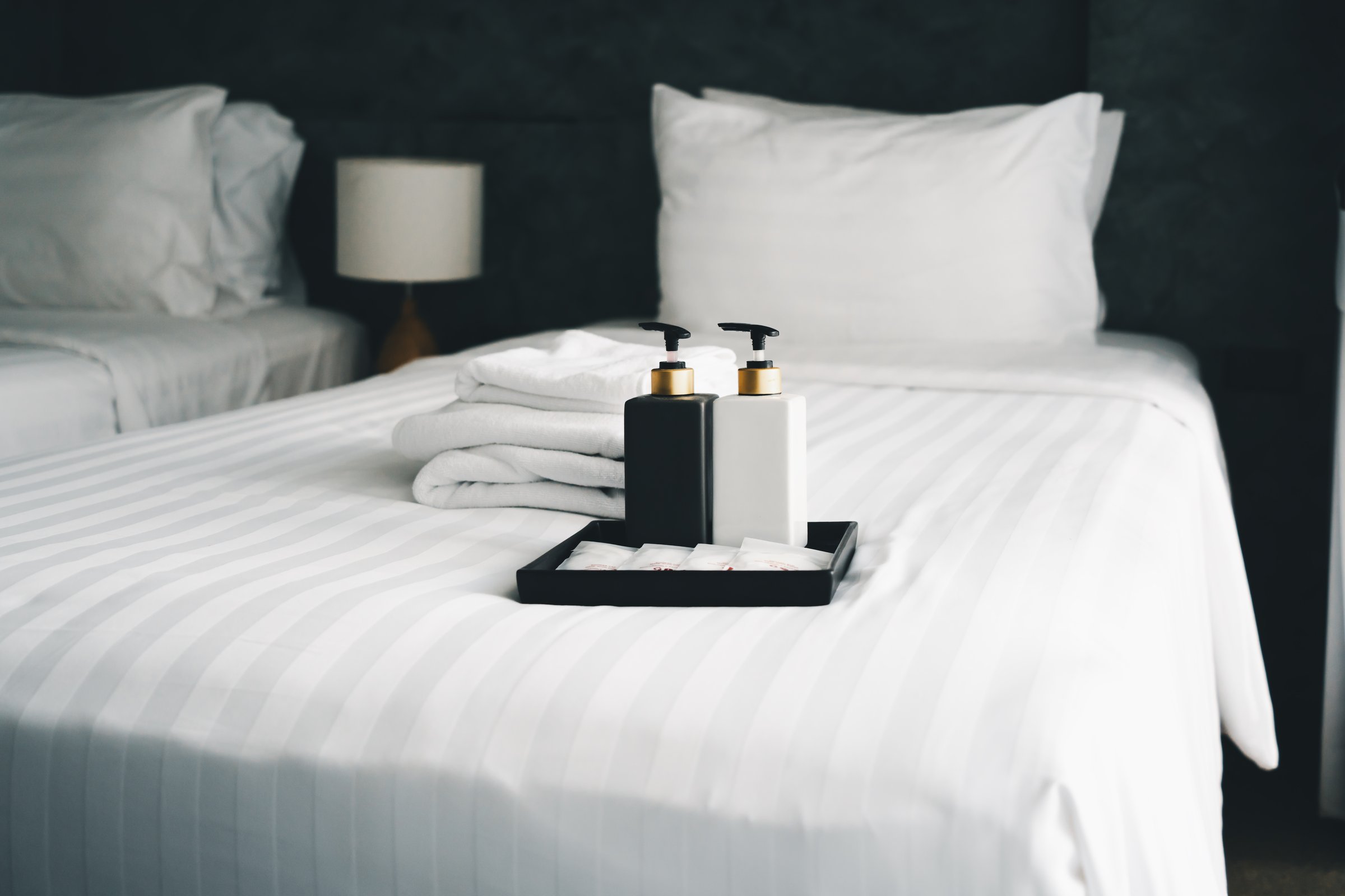 bottles of bathroom accessories with blurred towels on white cleaned bed in hotel bedroom, in soft focus