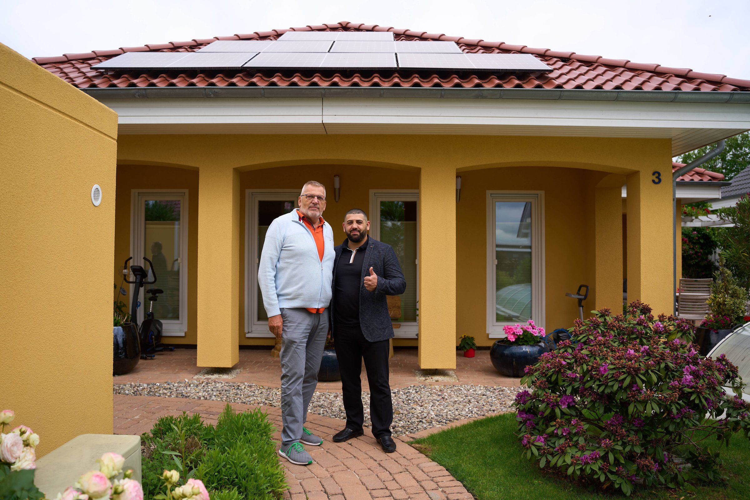 Two men standing outside a yellow house with solar panels on the roof, surrounded by shrubs and flowers.