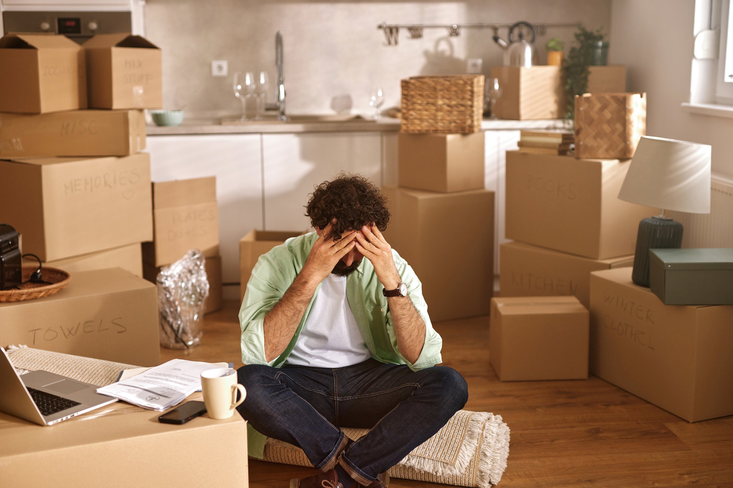 Stressed man sitting on the floor of his new apartment surrounded by moving boxes, holding his head in his hands