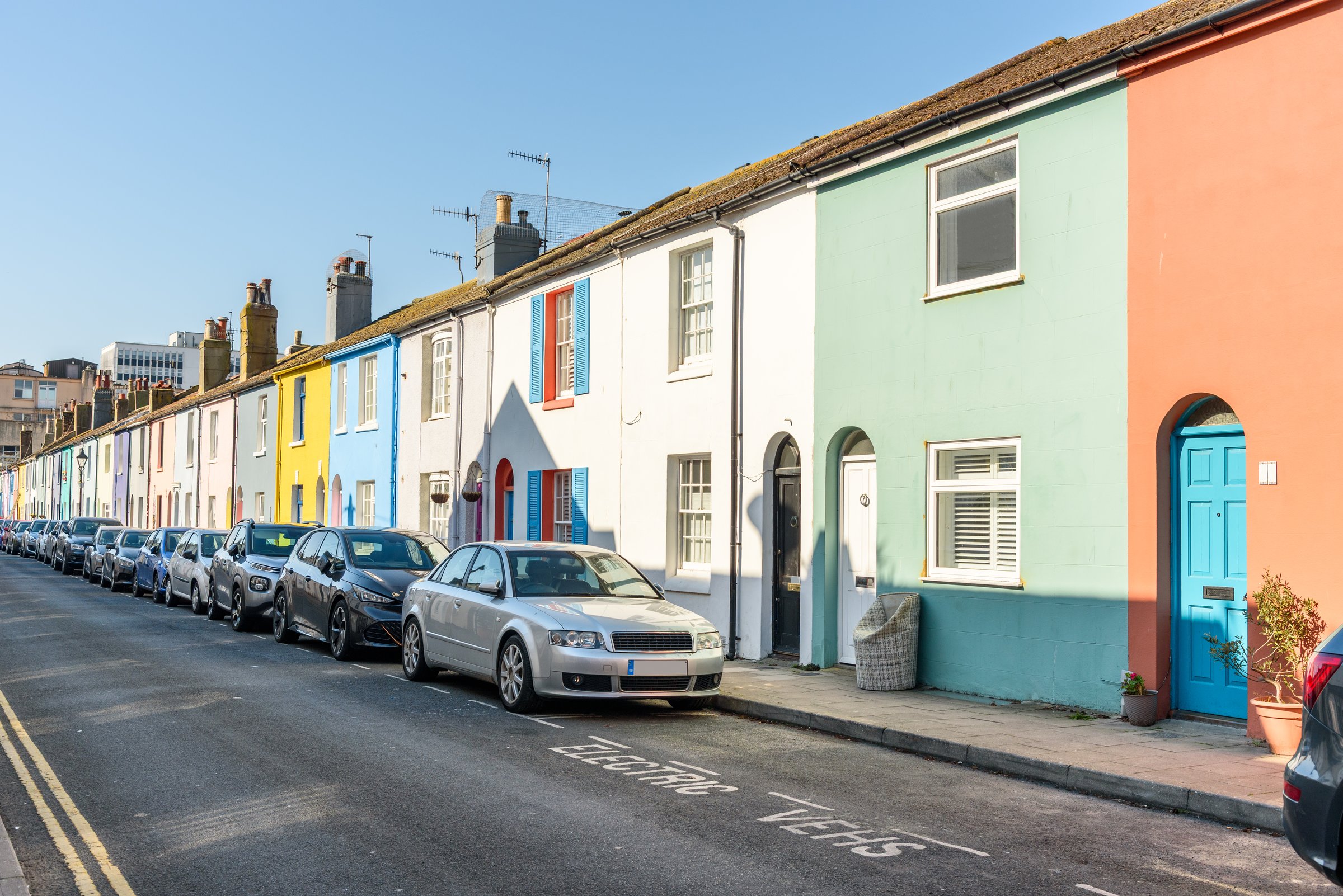 Row of colourful terraced houses along a street under clear sky in spring. An empty parking space for electric vehicles is in foreground. Brighton, England, UK.