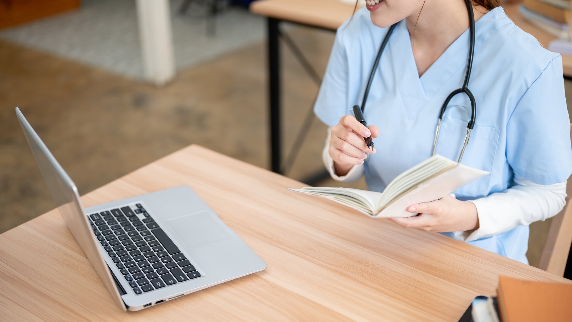 A close-up of a beautiful Asian female medical student in scrubs reading her lecture book while working on her research on a laptop in the library. medical school, college, education