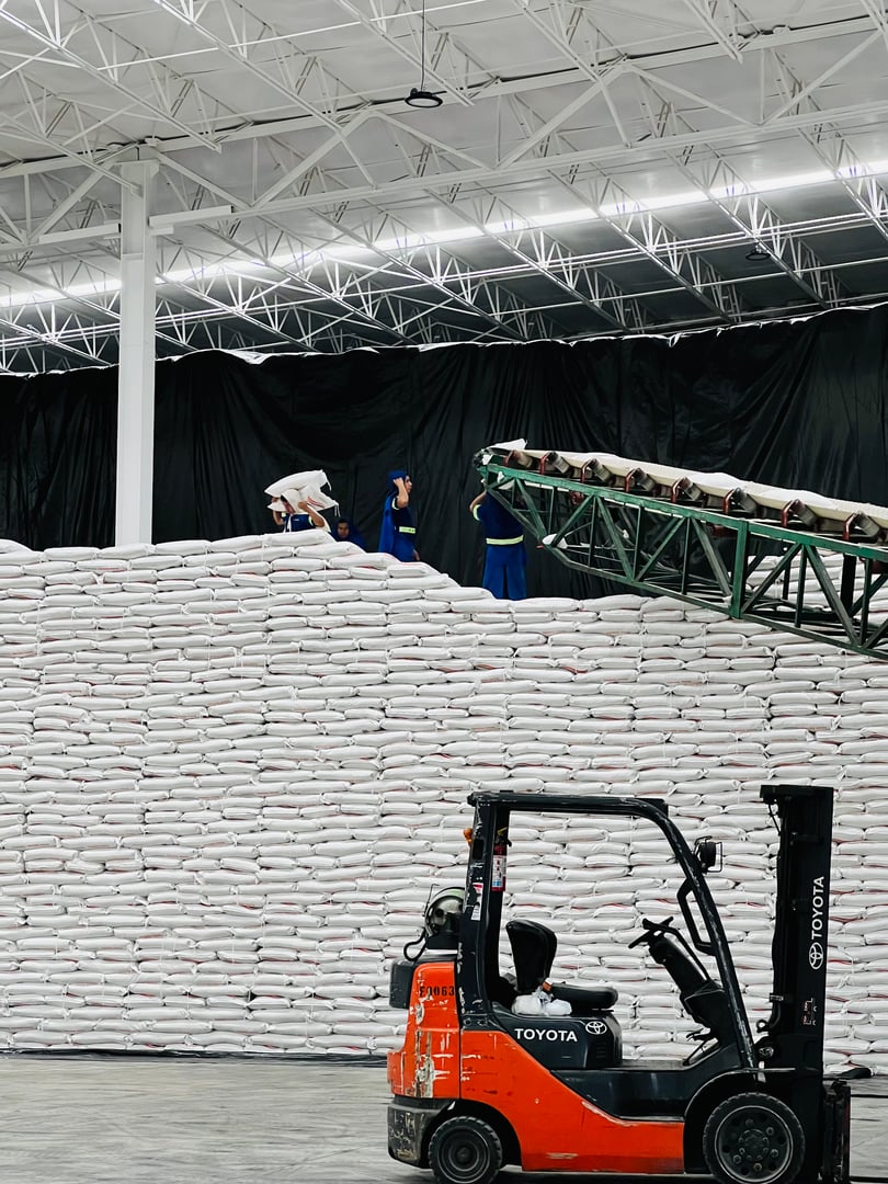 Workers in a warehouse stacking large bags onto a conveyor belt. A forklift is parked in the foreground.