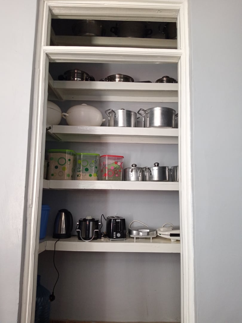 A pantry with shelves holding pots, kettles, food containers, and kitchen appliances against a light grey wall.