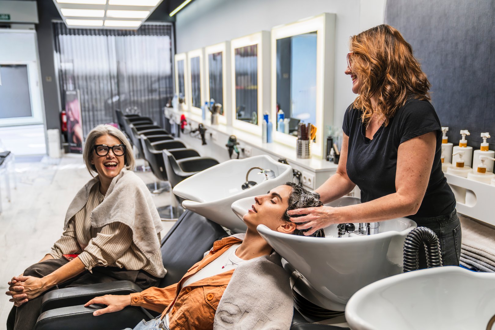 Stylist washing a client's hair in a salon while another customer waits, creating a happy social scene
