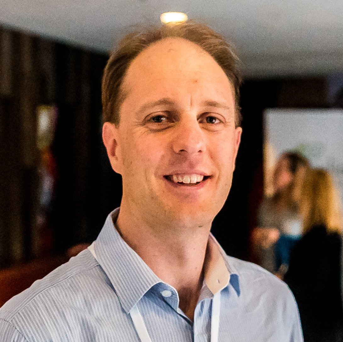 Smiling man in a striped shirt with a lanyard in a dimly lit indoor setting.