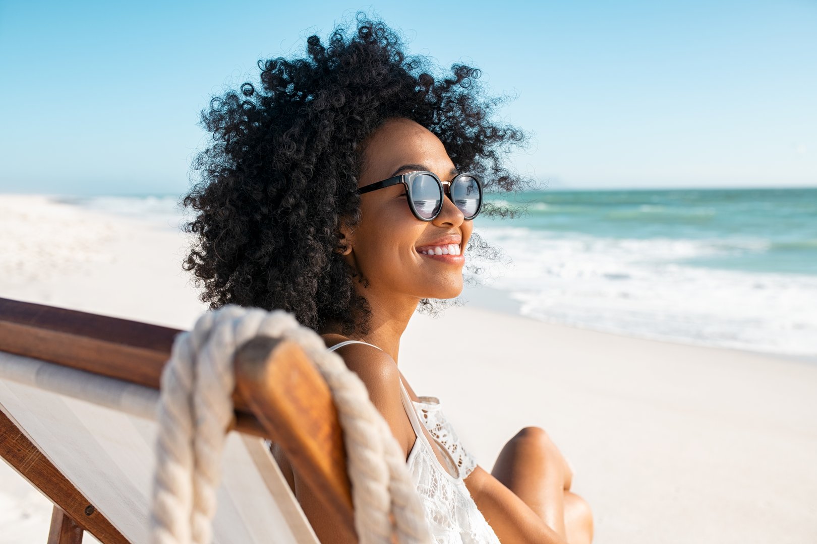 Portrait of happy young black woman relaxing on deck chair at beach wearing spectacles. Smiling african american girl with sunglasses enjoy vacation at beach. Carefree happy young woman sunbathing and relaxing at sea with copy space.