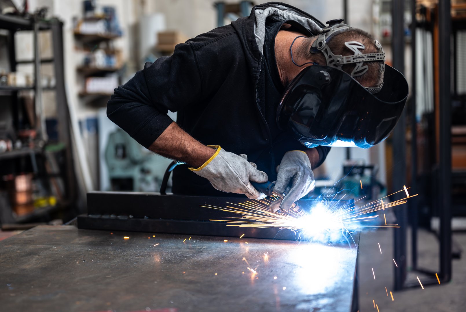 Performing a weld: the man in the welding mask is positioned with the welding machine on the welding point. When he activates it, a blue glow with smoke and orange sparks illuminates the scene.