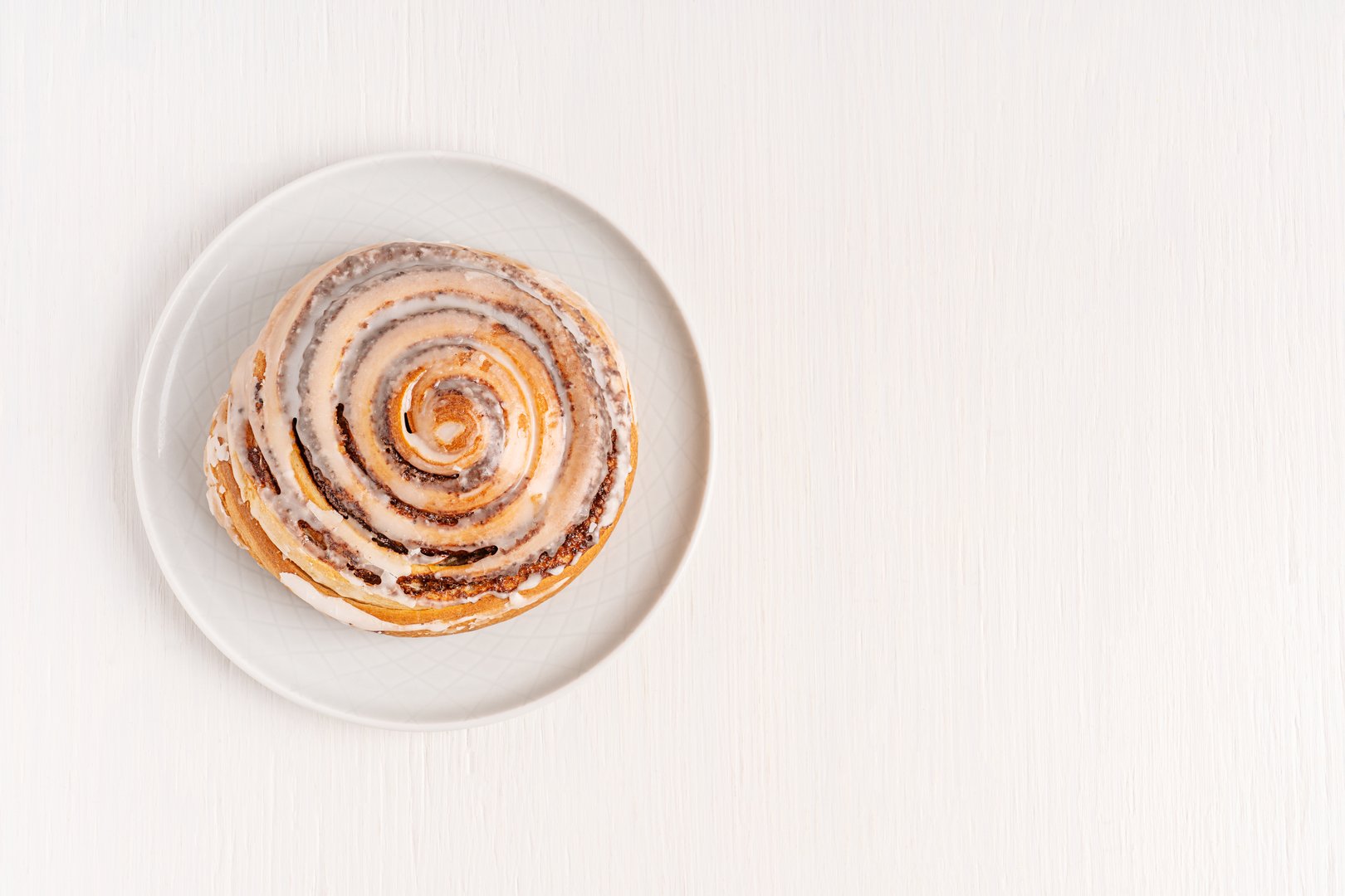 Top view of single sweet delicious homemade cinnamon roll bun or swirl glazed with sugar icing served on plate on white wooden background with copy space as comfort food dessert baked for breakfast