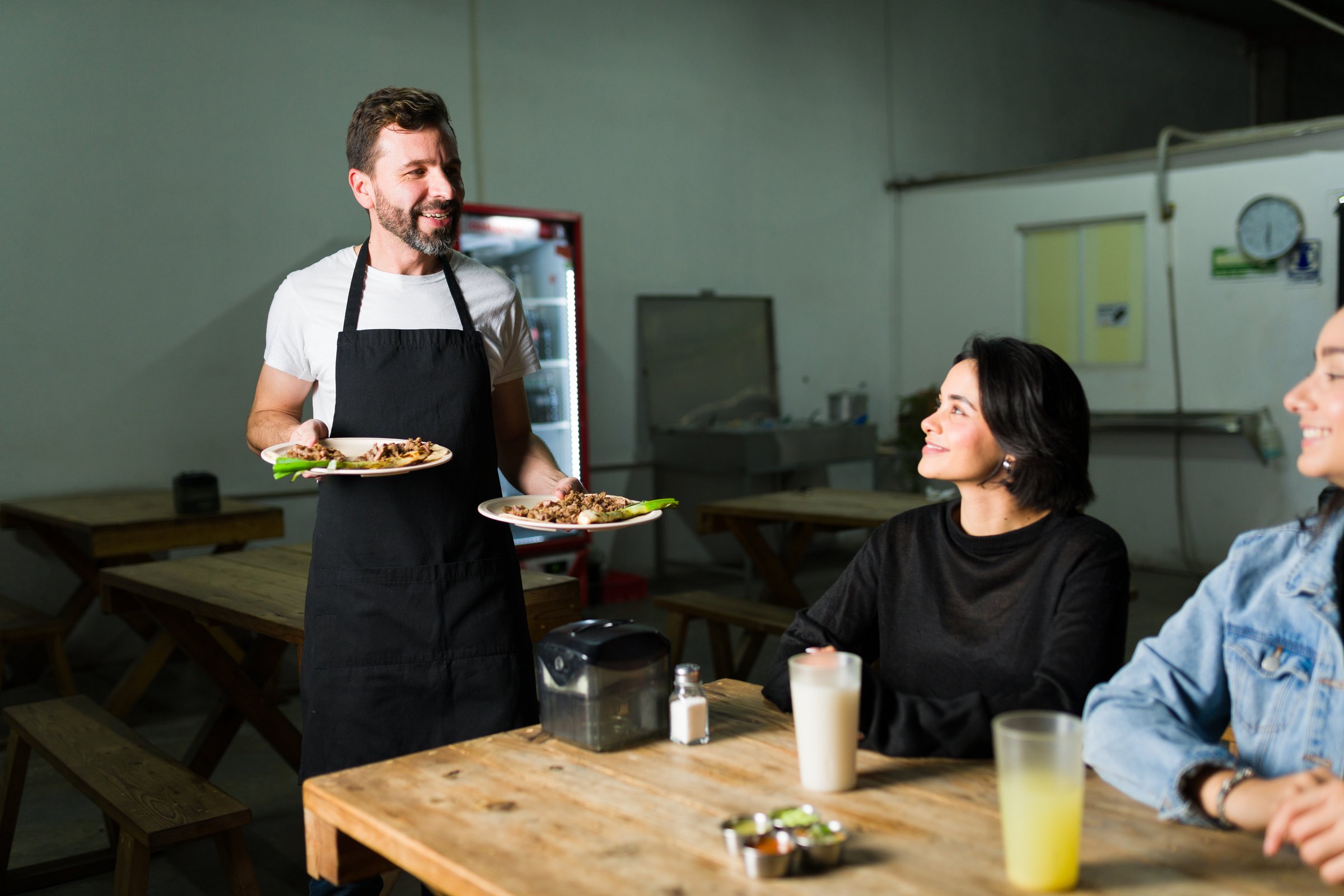 Happy waiter carrying two plates of delicious tacos to customers in a traditional mexican taqueria