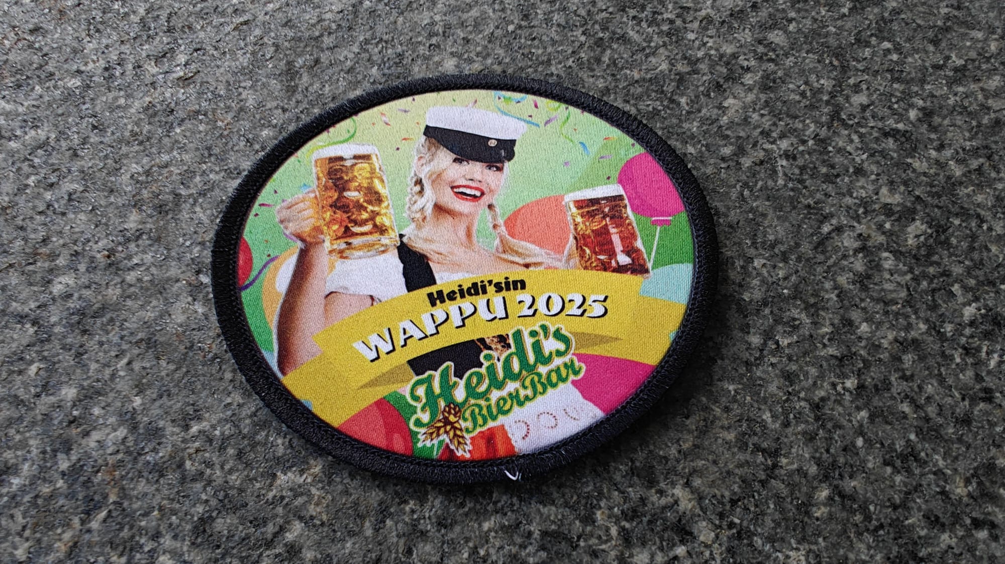 A round patch featuring a woman in traditional attire holding beer mugs, with text Heidis WAPPY 2025 Heidis Bier Bar.