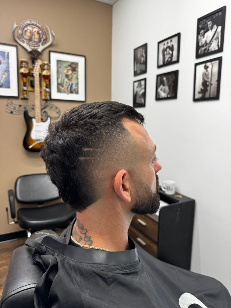 Man with short haircut and shaved design on side, sitting in barbershop with guitar and photos on the wall.