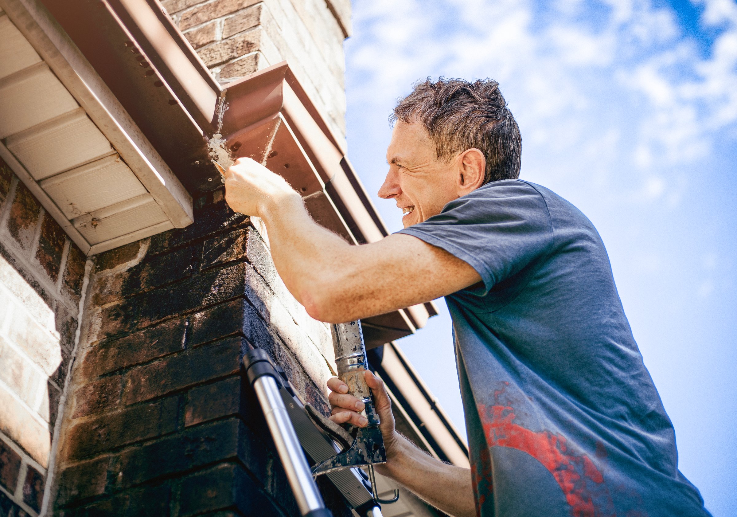 A man seals a gutter on the roof of a brick house overlooking a blue sky.
