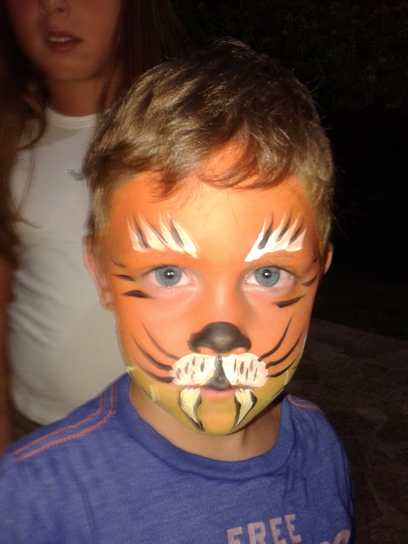 Child with lion face paint wearing a blue shirt, standing outdoors with another person partially visible in the background.