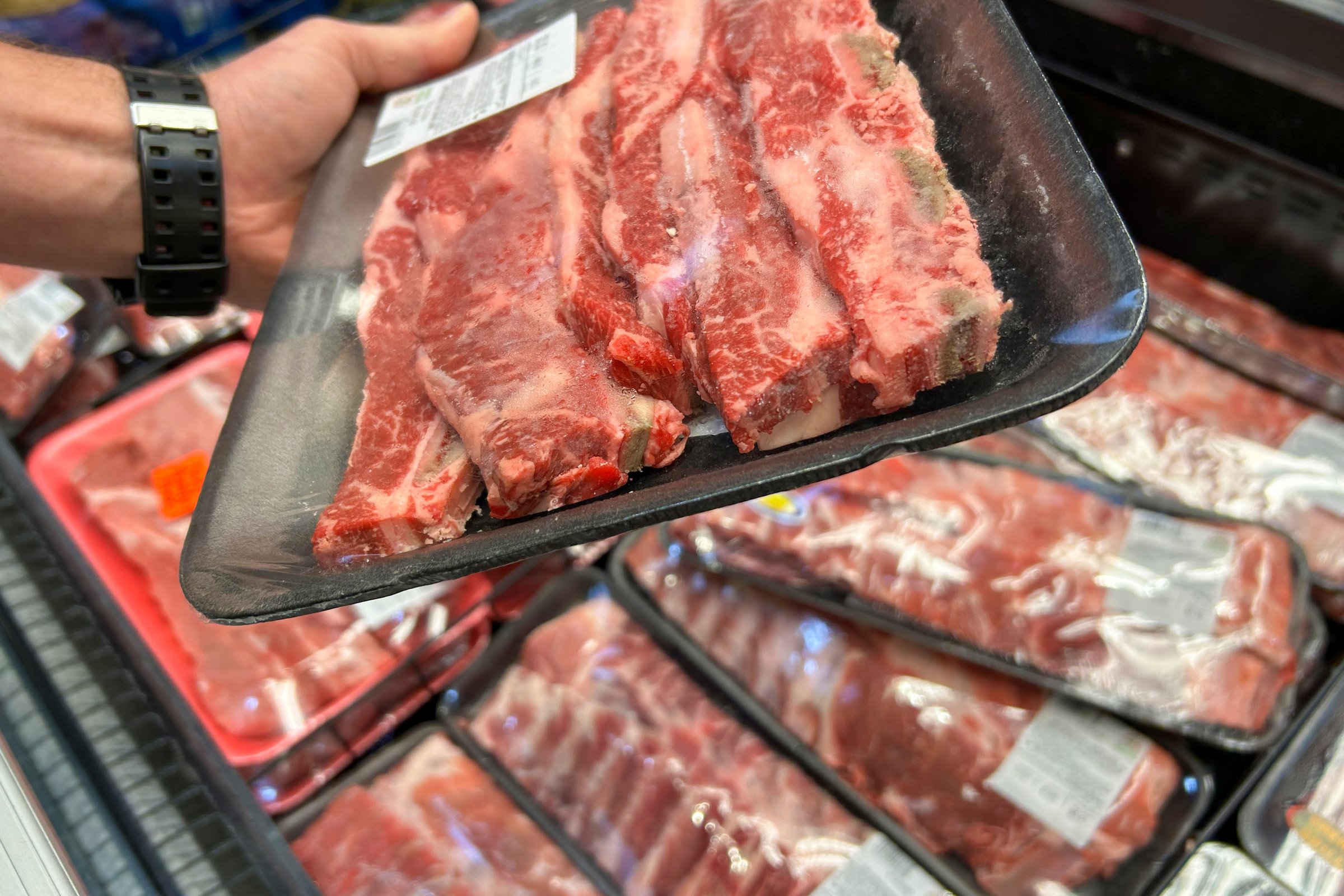 a person's hand holding a tray of packaged raw beef ribs, with more trays of meat in the background at a grocery store