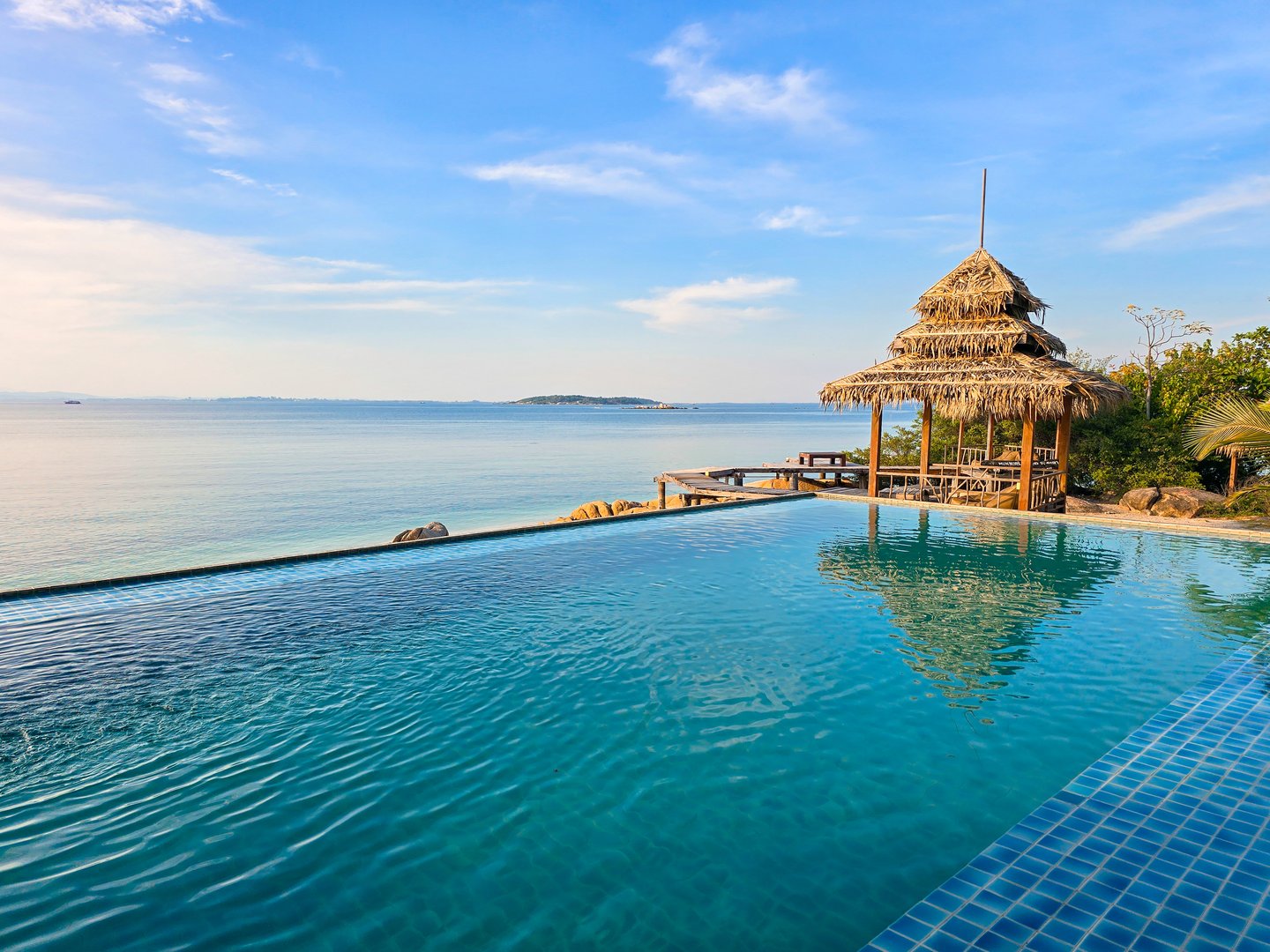 A luxurious infinity pool offers breathtaking views of the tranquil waters of Thailand. The sun begins to set behind the horizon, casting golden hues across the sky and water.