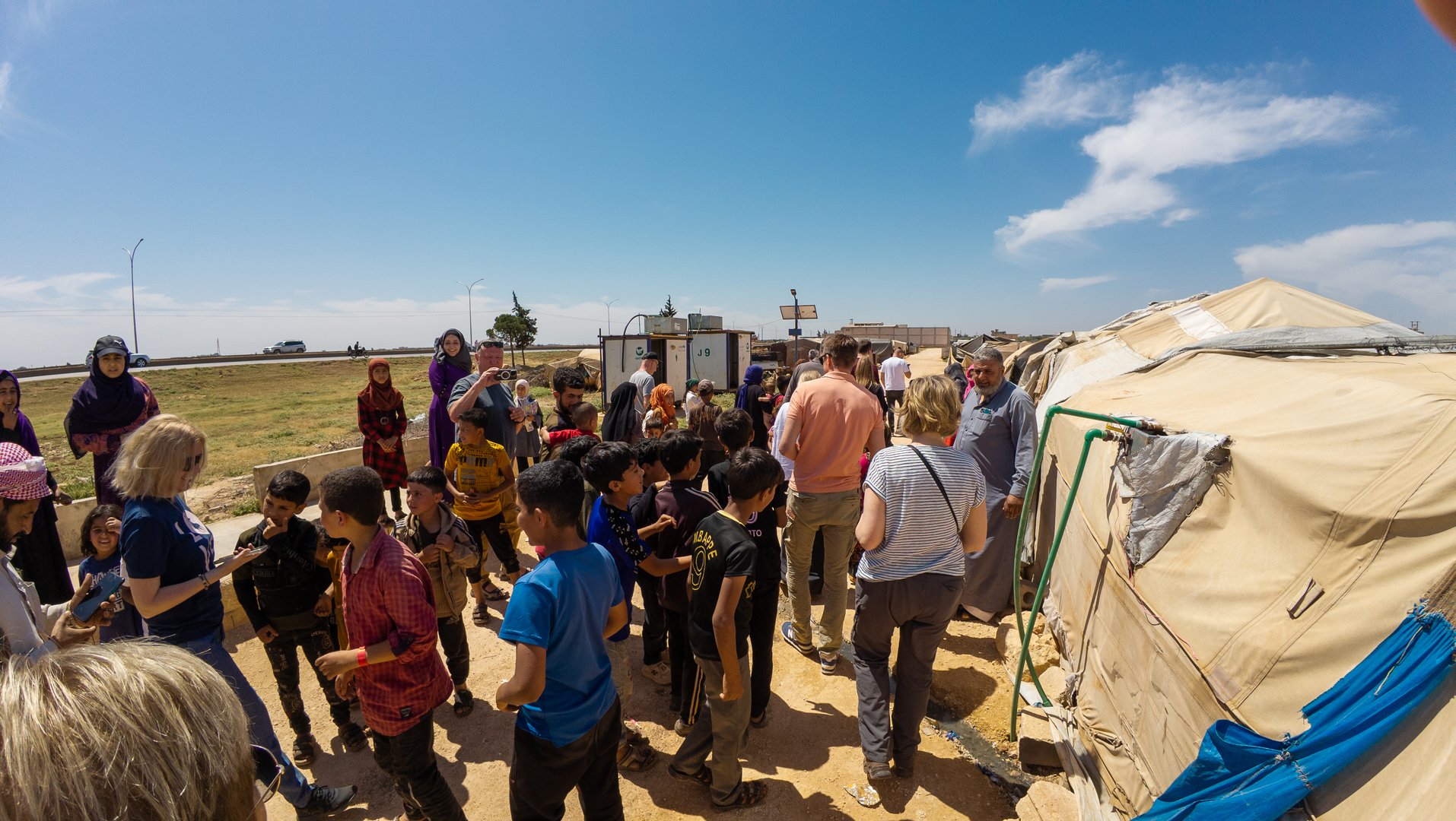 22/05/2025. Refuge camp near Aleppo, Syria. A group of people, including children and adults, gather near tents in an open field under a clear sky. A group of refuges meets visiting traveling helpers